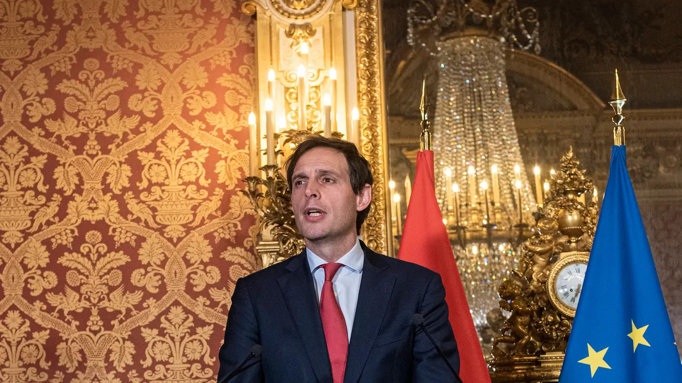 EU Commissioner Wopke Hoekstra speaking at an official event, with European and national flags in the background, discussing taxation and policy priorities.